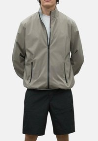 Chaqueta de cremallera gris claro con cuello alzado, bolsillos laterales y detalles de cremallera negra, combinada con pantalones cortos negros de diseño sencillo.