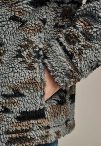 Fleece jacket in gray, brown, and black with a textured, shaggy surface. Features side pockets and a relaxed fit.
