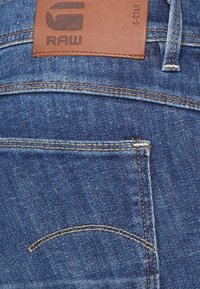 Jeans de mezclilla azul con un parche de cuero marrón etiquetado como "RAW". Notables detalles de costura y bolsillo acentúan el diseño clásico.