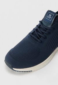 Chaussure de sport bleu marine avec une tige en tricot texturé, lacets plats, col rembourré et une semelle en caoutchouc légère en contraste. Étiquette de logo sur la languette.