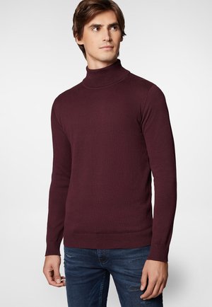 Burgundowa sweter z golfem, prążkowane mankiety i dół, dopasowany fason, miękka faktura, noszony z postrzępionymi niebieskimi dżinsami, model zwrócony nieco bokiem.