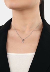 Collar de plata con una cadena, que presenta un pequeño colgante redondo y un acento ovalado, lucido sobre piel desnuda debajo de un blazer negro.