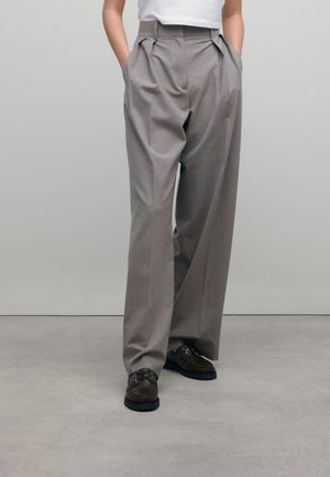 Persoon draagt high-waisted, wijde grijze broek met plooien, een ingestopt wit shirt en donkerbruine veterschoenen, staand op een grijze vloer.