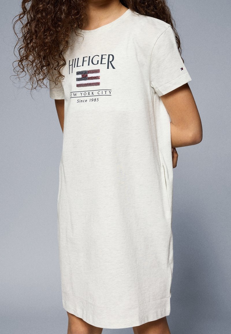 Světle béžové šaty ve formě trička vyrobené z měkké bavlny, s potiskem loga "HILFIGER", designem vlajky a krátkými rukávy s ohrnutými manžetami.