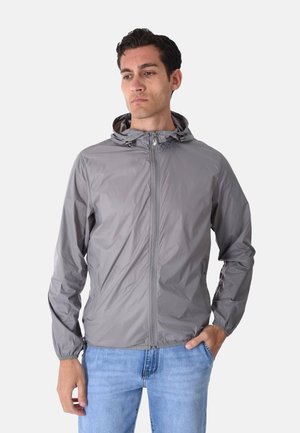 Uomo con giacca grigia con cappuccio e zip e jeans azzurri chiari, in piedi con una mano in tasca, guardando leggermente in basso e di lato.