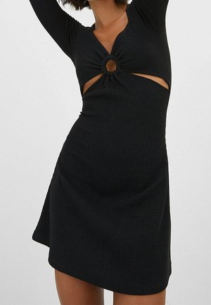 Vestido negro de canalé con mangas largas, que presenta un escote en V y detalles recortados en la cintura, acentuados por un elemento de metal circular.