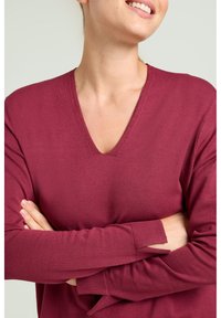 Bordeauxrode gebreide trui met V-hals, lange mouwen met uitsnijdingen en een gladde textuur, gedragen met gekruiste armen.