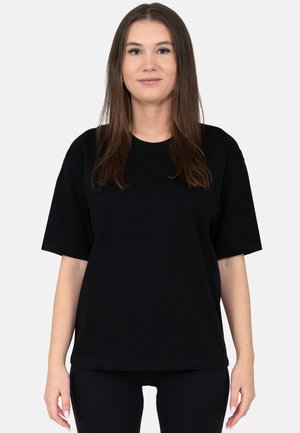 Frau mit glattem braunem Haar, die ein schlichtes schwarzes, übergroßes T-Shirt mit kurzen Ärmeln trägt und vor einem hellgrauen Hintergrund steht.