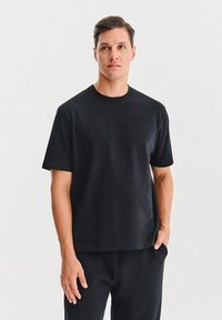 Mężczyzna z krótkimi brązowymi włosami w prostym czarnym T-shircie i czarnych spodniach stoi z jedną ręką w kieszeni na białym tle.