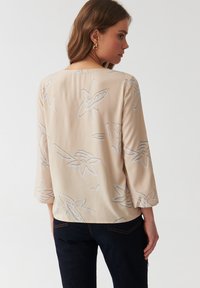Beige Bluse mit drei Viertelarmen, die ein florales Muster in Hellgrau aufweist. Glatte Textur und lockere Passform, getragen mit dunklen Jeans.