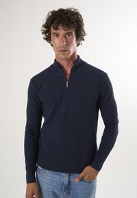 Marine blauwe ritssweater met geribbelde textuur, opstaande kraag en lange mouwen, gecombineerd met lichtblauwe spijkerbroek. Minimalistisch ontwerp, zonder patronen.