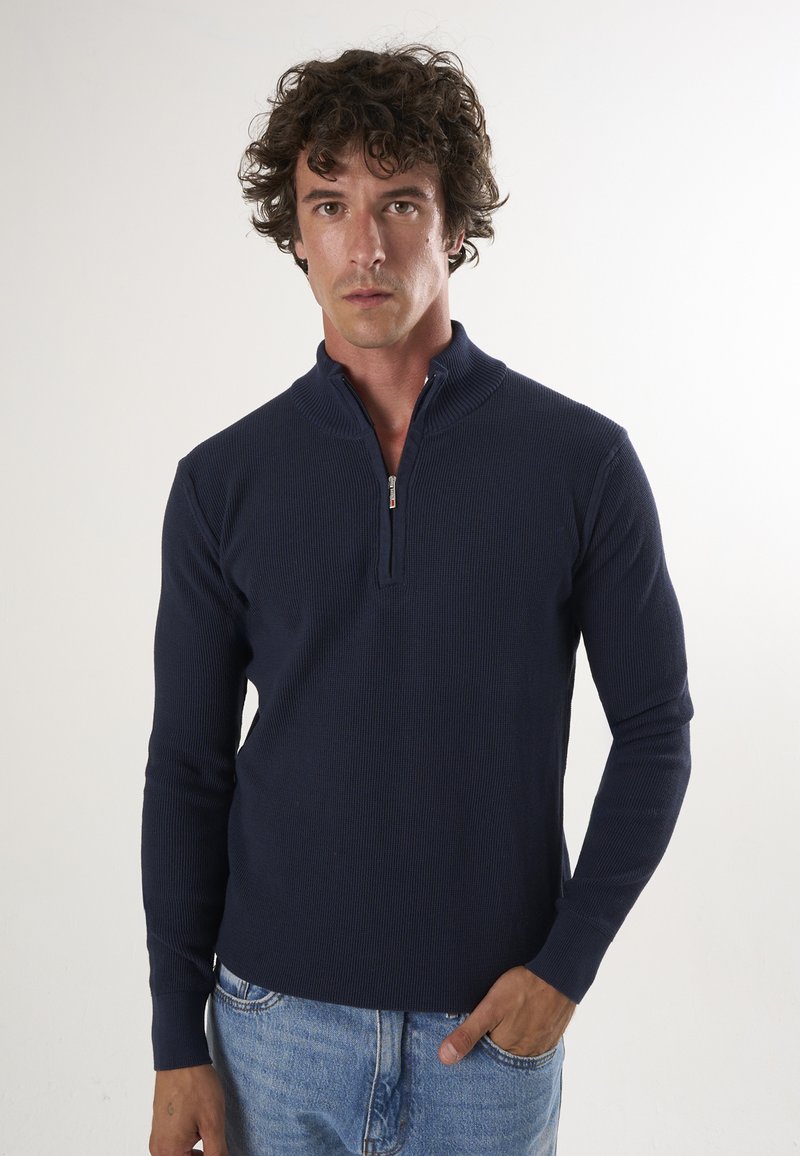 Marine blauwe ritssweater met geribbelde textuur, opstaande kraag en lange mouwen, gecombineerd met lichtblauwe spijkerbroek. Minimalistisch ontwerp, zonder patronen.