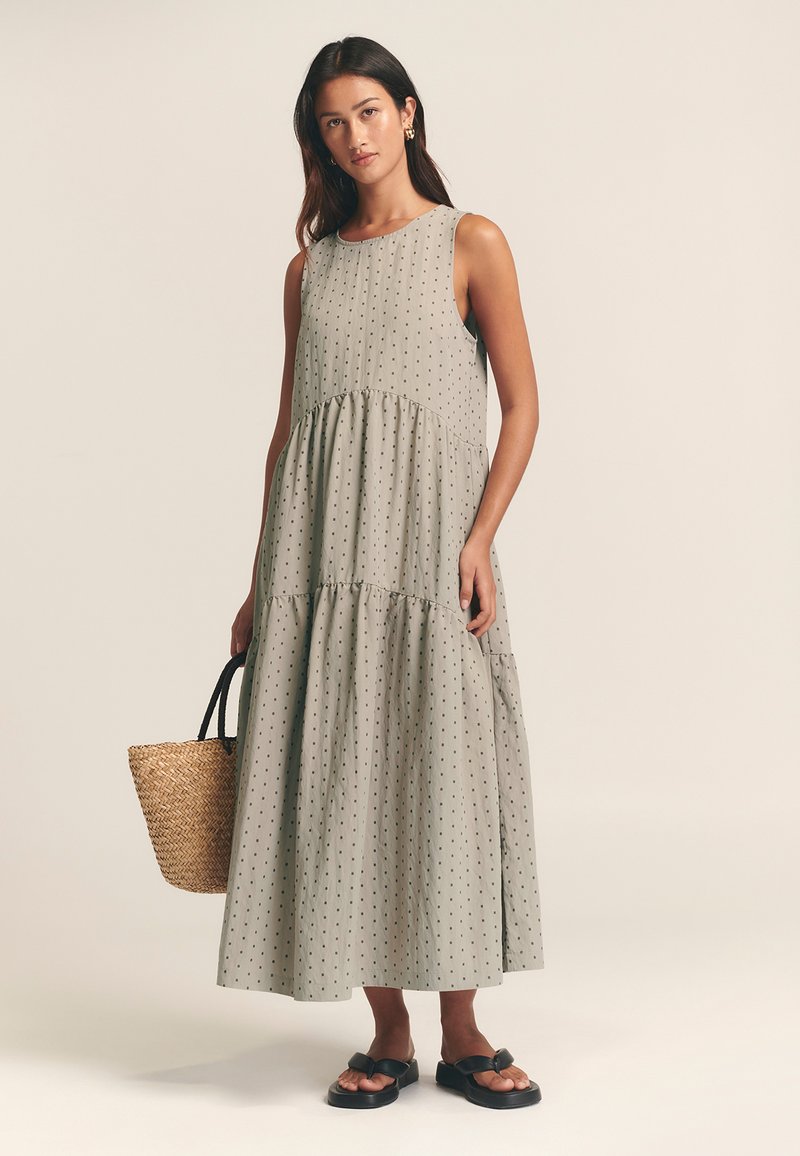 Svetlo zelena večplastna maxi obleka z majhnimi črnimi pikami, z okroglim izrezom, brez rokavov, v kombinaciji z vzorčasto torbo in črnimi sandali.