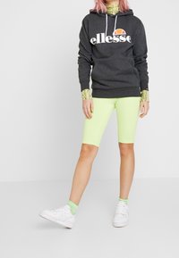 Sudadera gris oscuro con el logo blanco de 'ellesse', combinada con pantalones cortos de ciclista verde lima. Zapatillas blancas y mangas con estampado completan el atuendo.