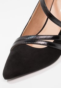 Escarpin noir à bout pointu en matière suédée, avec des brides croisées noires brillantes et une doublure intérieure lisse.