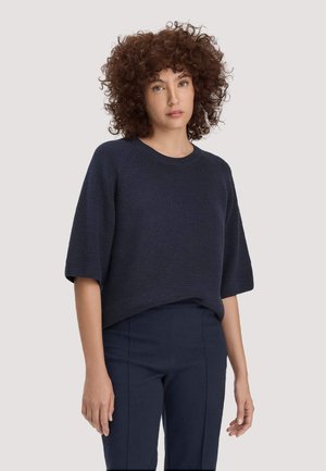 Frau mit lockigem braunem Haar, die einen marineblauen kurzärmeligen Strickpullover und passende marineblaue, eng geschnittene Hosen vor einem einfarbigen Hintergrund trägt.