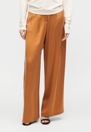 Ženska v hlačah z visokim pasom in širokimi hlačnicami rjave rjave barve z gubami spredaj ter črnimi sandali z odprtimi prsti, na enobarvnem ozadju.