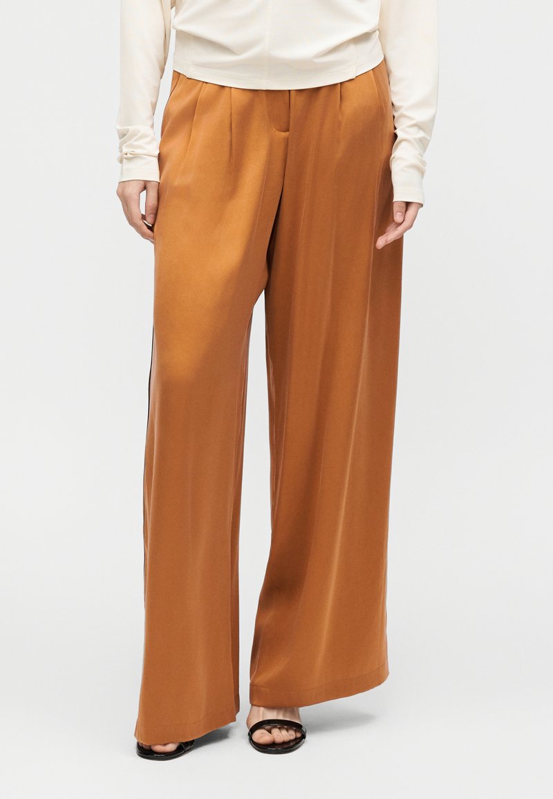 Γυναίκα που φοράει παντελόνι ψηλόμεσο, φαρδύ με πιέτες μπροστά, σε σκουριά απόχρωση, και μαύρα ανοιχτά σανδάλια, μπροστά σε απλό φόντο.