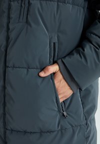 Mørkeblå isoleret jakke med en hånd i sidelommen, der har en quiltet tekstur og en ribbet manchet ved håndleddet.