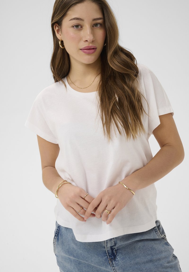 Jeune femme aux longs cheveux bruns portant un t-shirt blanc, un jean bleu et des bijoux en or, notamment des boucles d'oreilles créoles, des bracelets, un collier et des bagues.