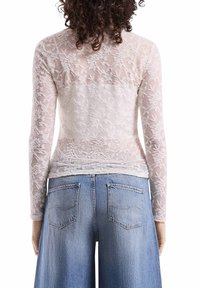 Langärmliges, transparentes weißes Oberteil mit floralem Spitzenmuster und glatter Rückseite, kombiniert mit hellblauen, weit geschnittenen Jeans mit Gesäßtaschen.