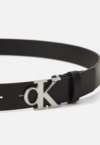 Mustkas nahkrihm, millel on hõbedane metallklamber "CK" logo. Sujuv tekstuur ja lihtne, klassikaline disain, millel on reguleeritavad augud.