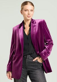 Blazer de terciopelo púrpura con cierre de un solo botón, solapas con muesca y bolsillos delanteros; combinado con una blusa negra y jeans grises.