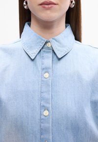 Világoskék farmering klasszikus gallérral, gombos elejével és puha textúrájával. Off-white gombokkal és diszkrét varrási részletekkel rendelkezik.