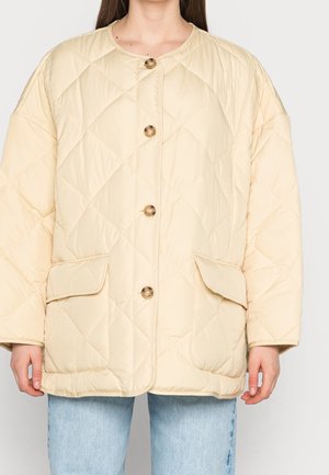 Femme portant une veste matelassée beige avec de gros boutons et deux poches à rabat sur le devant, associée à un jean bleu clair.