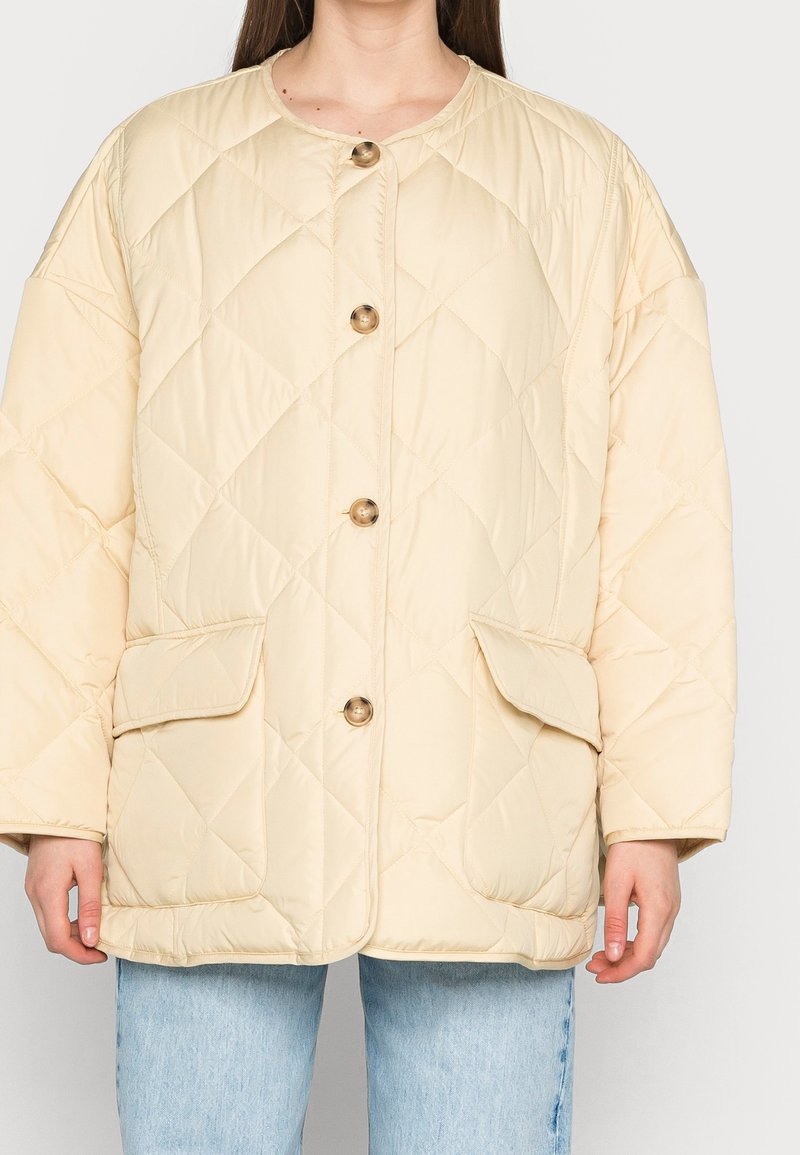 Femme portant une veste matelassée beige avec de gros boutons et deux poches à rabat sur le devant, associée à un jean bleu clair.