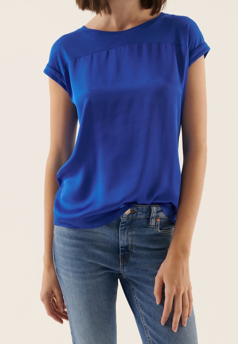 Femme portant un chemisier bleu vif à manches courtes, légèrement rentré dans un jean denim bleu clair, sur un fond clair uni.