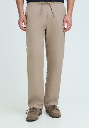 Hombre vistiendo pantalones beige con cordón y zapatos marrones con cordones, de pie frente a un fondo claro uniforme.