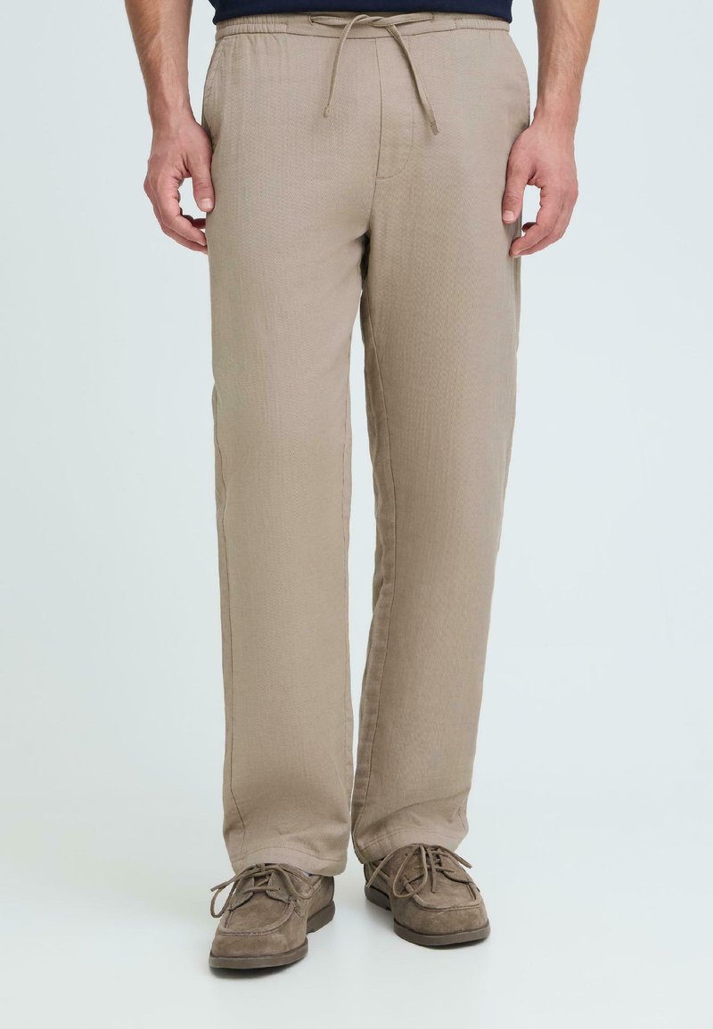 Άνδρας που φοράει μπεζ παντελόνι με κορδόνι και καφέ παπούτσια δεμένα με κορδόνια, στέκεται μπροστά σε απλό φωτεινό φόντο.