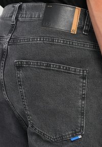 Vaqueros de mezclilla gris con una superficie texturizada, bolsillos traseros, costuras en contraste y una etiqueta negra con acento marrón en la cinturilla.