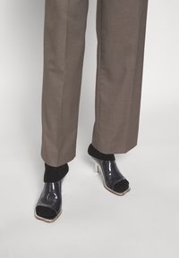 Klart högklackade skor med en genomskinlig överdel och svart texturerad tåöverdrag, kombinerat med bruna skräddarsydda byxor och svarta strumpor.