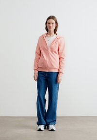 Γυναίκα που στέκεται φορώντας ροζ ανοιχτό φούτερ με φερμουάρ, λευκό μπλουζάκι, μπλε τζιν και λευκά αθλητικά παπούτσια σε απλό φωτεινό φόντο.