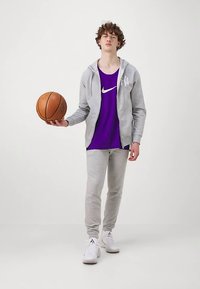 Pelēks kapuci, purpursarkans Nike bez Piedēkļa krekls un pelēki sporta bikses ar baltām sneakers. Turēšana basketbola bumbā. Gluds audums, brīvs piegriezums, ikdienas sporta apģērbs.