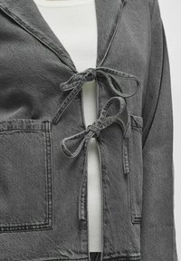 Šedá riflová bunda so šálovým golierom, zaviazaná vpredu, s dvoma prednými vreckami a textúrovanou tkaninou. Biele vnútorné vrstvy sú viditeľné pod ňou.