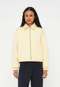 Dzeltena polsterēta bomberjakete ar rāvējslēdzēju, ribotām piedurknēm un apkakli. Uz krūtīm ir izšūts logs. Gluda tekstūra.