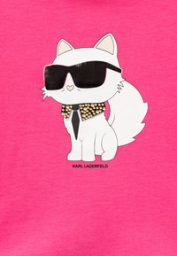 Chemise rose à manches courtes mettant en avant un chat cartoon avec un pelage blanc, des lunettes de soleil noires et un collier à clous. Nom de la marque affiché sous le design.