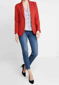 Donna con un blazer rosso, una blusa a fantasia, jeans blu e scarpe col tacco nere, in piedi davanti a uno sfondo bianco.