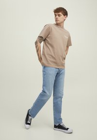 Beige short-sleeve t-shirt with white text, paired with light blue jeans and black sneakers with white accents. Casual fit and style.