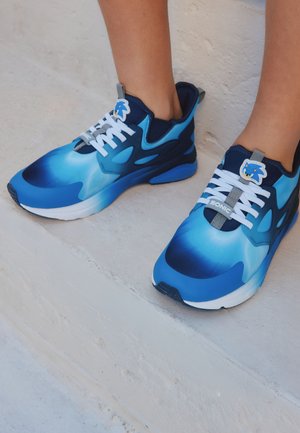 Niebiesko-białe sneakersy z logo Sonic the Hedgehog, noszone na bose stopy, stojące na białych teksturowanych stopniach.