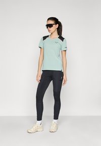 Lys grøn treningstrøye med svarte og røde detaljer, kombinert med svarte leggings med teksturerte paneler og kremfargede treningssko.
