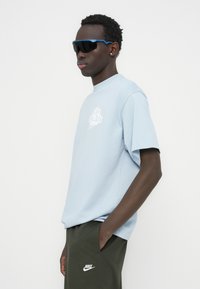 Ανοιχτό μπλε κοντομάνικο μπλουζάκι με το λευκό γραφικό «Live On Air»· συνδυασμένο με σκούρο πράσινο παντελόνι με το λογότυπο της Nike. Μαύρα γυαλιά ηλίου με μπλε λεπτομέρειες.