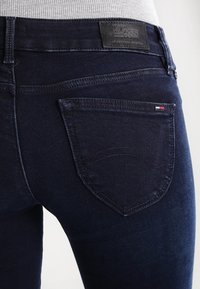 Mörkblå denimjeans med en slät textur, som har en distinkt bakfickedesign och en läderlogotyp på midjebandet.