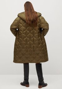 Olivengrøn quiltet jakke med hætte, med mønster af cirkulære former, knælængde snit og sorte knæhøje støvler.
