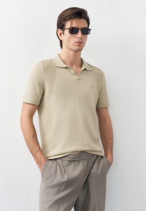 Homme portant un polo beige à manches courtes, un pantalon gris et des lunettes de soleil noires, debout devant un fond blanc uni.