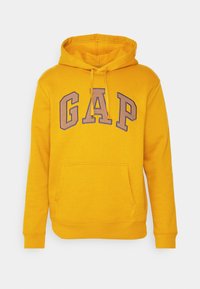 Sudadera amarilla con un bolsillo frontal, capucha con cordón y logo "GAP" en morado. La tela parece suave, con puños y dobladillo acanalados.