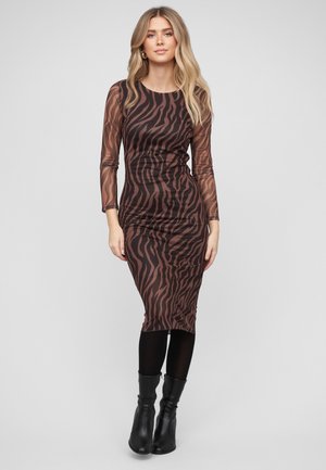 Kvinde med blondt hår iført en tætsiddende, langærmet midi-kjole med brune og sorte zebra-striber, sorte strømpebukser og sorte ankelstøvler.